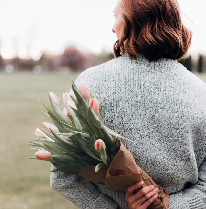 haircare concept - middle-aged woman with short red hair facing open field holding a bouquet of flowers behind her