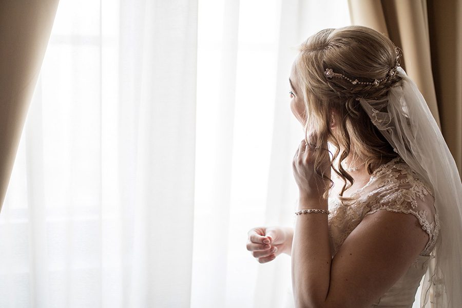 wedding hairstyle concept - blonde woman in bridal gown and wedding updo with veil looks out a window with sheer curtains